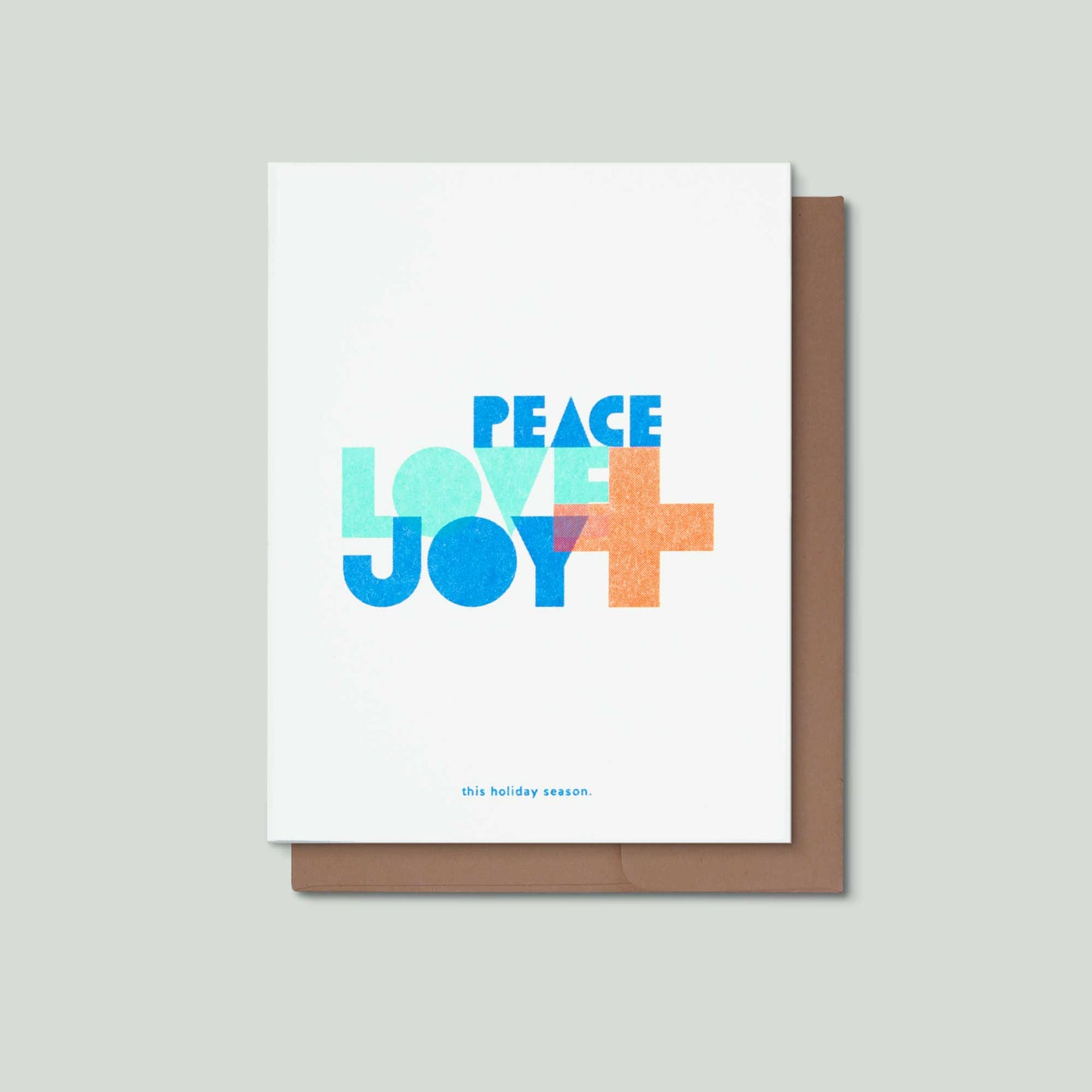An illustrated typographic risograph greeting card that says Peace, Love + Joy in the middle in large bold colorul font, and this holiday season small at the bottom. It is front of a kraft brown envelope on a grey background.