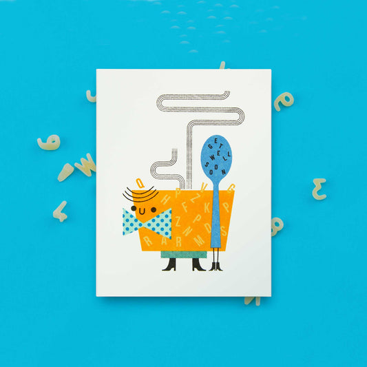 A risograph greeting card featuring an illustrated smiling bowl of alphabet soup, next a spoon with the words "get well soon" spelled out on it. It is in front of a blue backgound with dried alphabet pasta scattered around it.