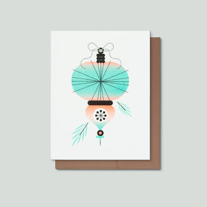 A greeting card featuring an illustrated Christmas ornament in front of a brown envelope and a grey background.