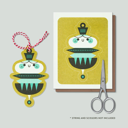 A risograph printed illustrated greeting card of an ornament with cut lines so that they ornament can be cut out of the card and hung. There is a sample of the cut out ornament with a red and white string and scissors.They are all on a grey background.