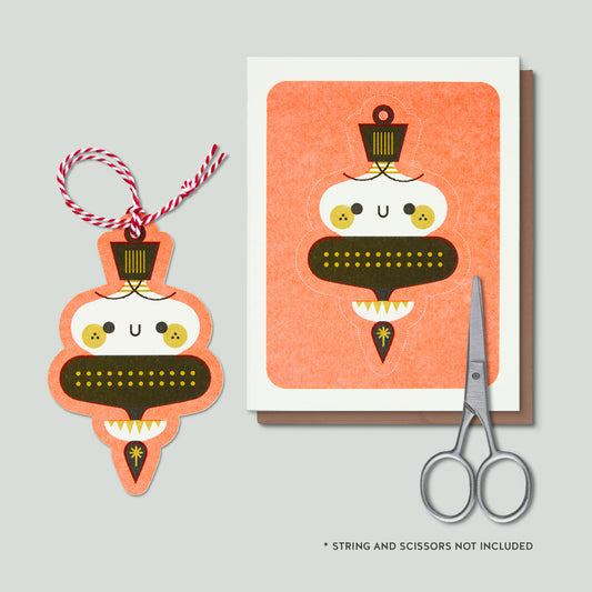 A decorative ornament shown beside a matching card, with a pair of scissors and small text noting that the string and scissors are not included. The ornament and card feature a smiling illustrated Christmas ornament on a light gray background.
