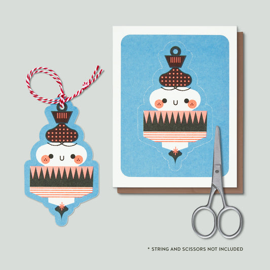 A product photo showing an ornament with a red and white string and a greeting card featuring the same illustration. A pair of silver scissors is placed beside the card, and small text at the bottom reads '* STRING AND SCISSORS NOT INCLUDED'.