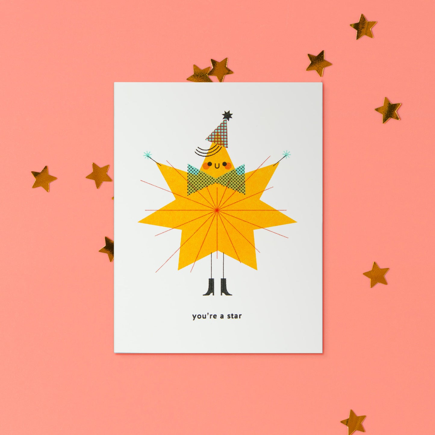 A white greeting card featuring a whimsical illustration of a star-shaped figure wearing a party hat and bow tie, with the text 'you're a star' at the bottom, displayed on a coral pink background with scattered gold star decorations.