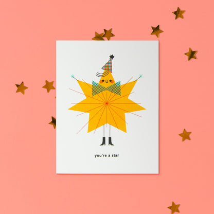 A white greeting card featuring a whimsical illustration of a star-shaped figure wearing a party hat and bow tie, with the text 'you're a star' at the bottom, displayed on a coral pink background with scattered gold star decorations.