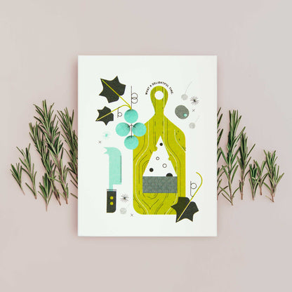 An illustrated greeting card featuring a stylized illustration of a green cutting board, grapes, leaves, and cheese, displayed on beige background with sprigs of rosemary