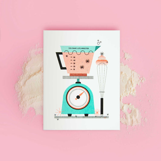 An illustrated risograph greeting card featuring a kitchen scale, measuring cup, and whisk displayed on a pink background with flour scattered over it.
