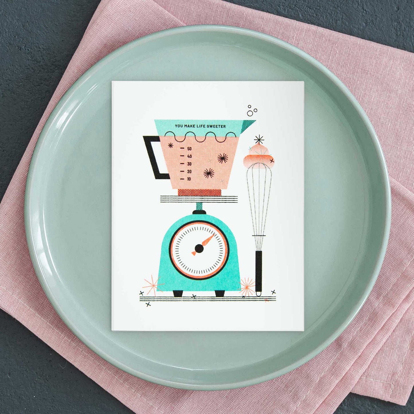An illustrated risograph greeting card featuring a kitchen scale, measuring cup, and whisk displayed on a light green ceramic plate over a pink textured cloth.
