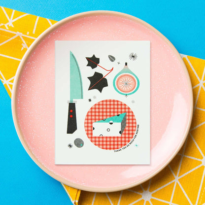 An illustrated risograph greeting card featuring illustrations of kitchen items and food, displayed on a speckled pink plate over a bright blue and yellow patterned surface.