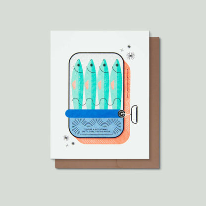 A greeting card with a whimsical illustration of four fish inside a can, shown with a brown envelope on a light gray background. The card features the text 'YOU"RE A BIT STINKY, BUT I LOVE YOU SO MUCH."