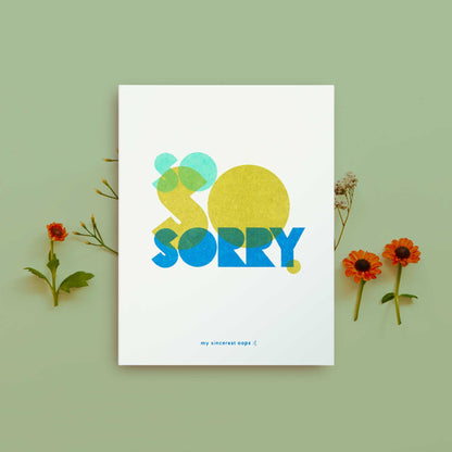 A white greeting card featuring a colorful graphic design that spells 'SO SORRY' using abstract shapes with 'my sincerest oops :(' at the bottom, styled flat lay on a sage green background with small orange and red flowers.