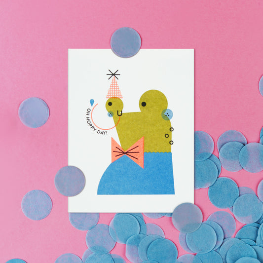 Illustrated card with a green frog with a party hat on a pink background with blue confetti