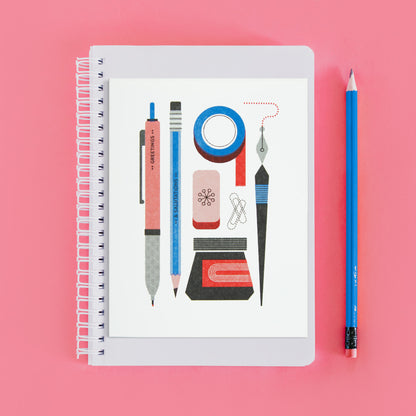 Notebook with a risograph printed greeting card sitting on top of it. The card has red, blue, and black illustrations of a pen, pencil, ink, tape, and an eraser on it. There is a blue pencil to the side, and it is on a pink backdrop.